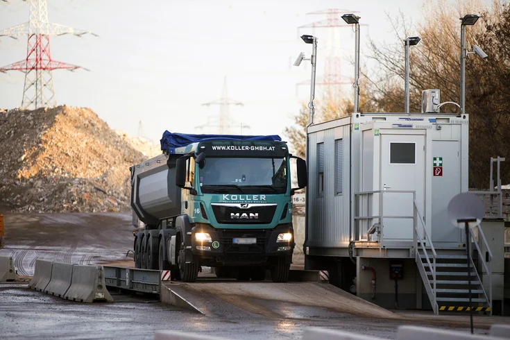 Foto: Lastwagen der Koller GmbH bei der Einfahrt ins Recycling Center Himberg auf der Waage; daneben Bürocontainer mit teilweise geschlossenen Jalousien, Türe und Scheinwerfern am Dach, im Hintergrund Schuttberge und Strommasten